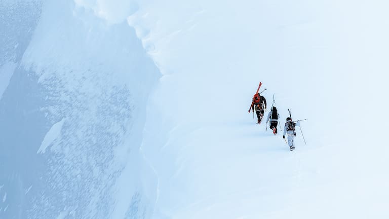 3 Personen steigen auf einen schneebedeckten Berg mit den Ski auf den Rücken und Houdini Sportswear Ausrüstung.