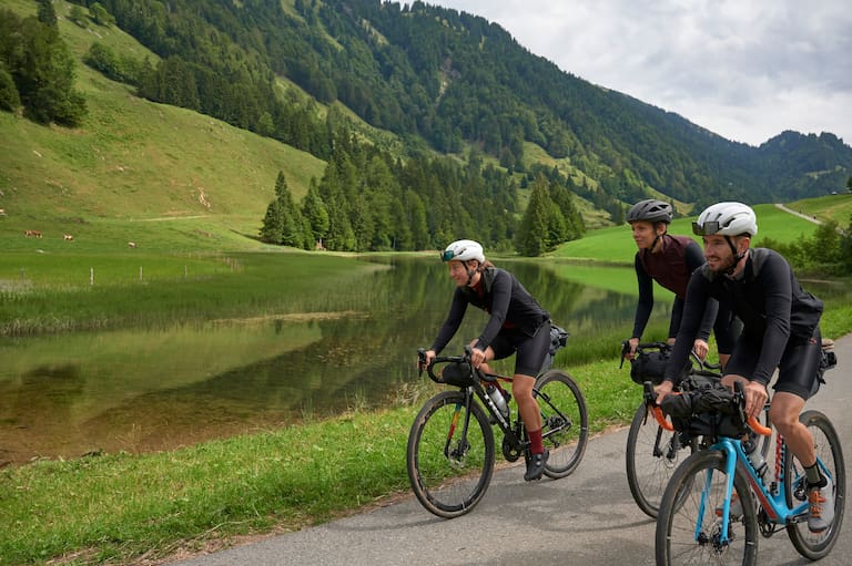 Drei Bikepacker mit leichtem Gepäck auf Gravel Bikes neben einem See und grünen Bergen im Bregenzerwald