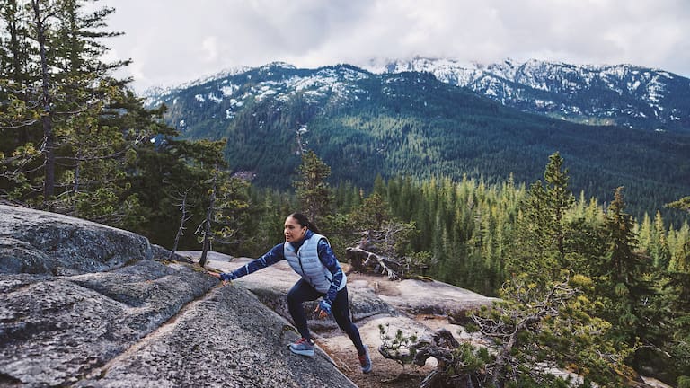 Eine Frau klettert in der Wildnis mit Outdoor-Bekleidung einen Hügel rauf.