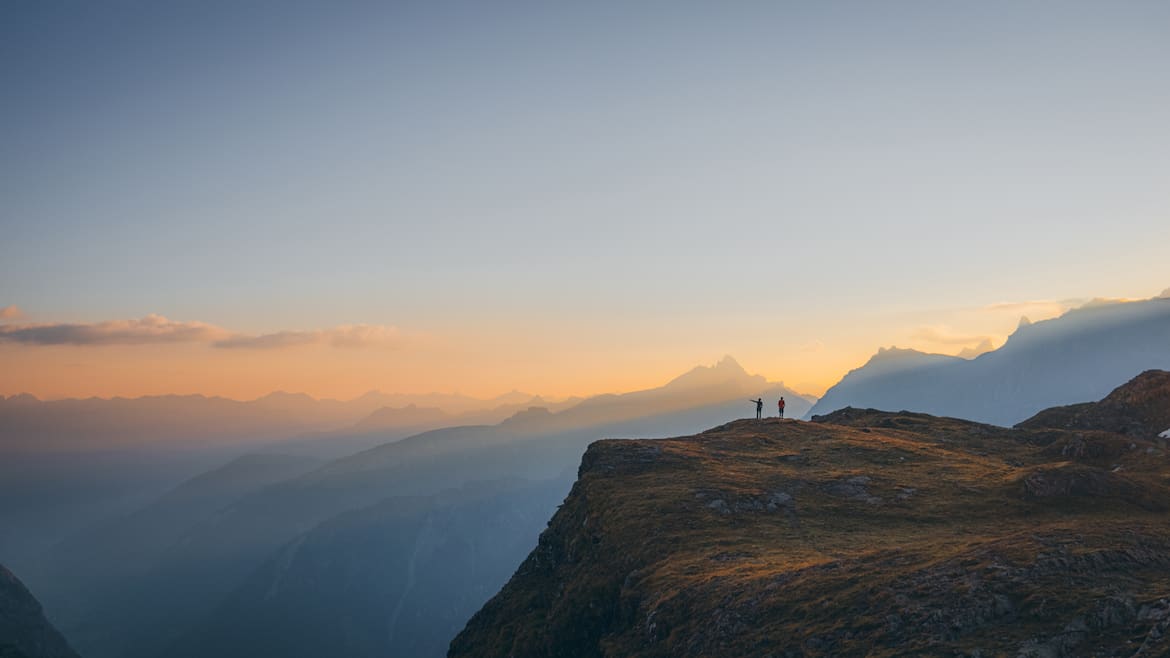 Kontrastreich: Layer für Layer präsentieren sich die Gipfel der Glarner Alpen im goldenen Licht der aufgehenden Sonne. Vor dem Horizont heben sich die zwei Personen am Berggipfel kontrastreich ab.